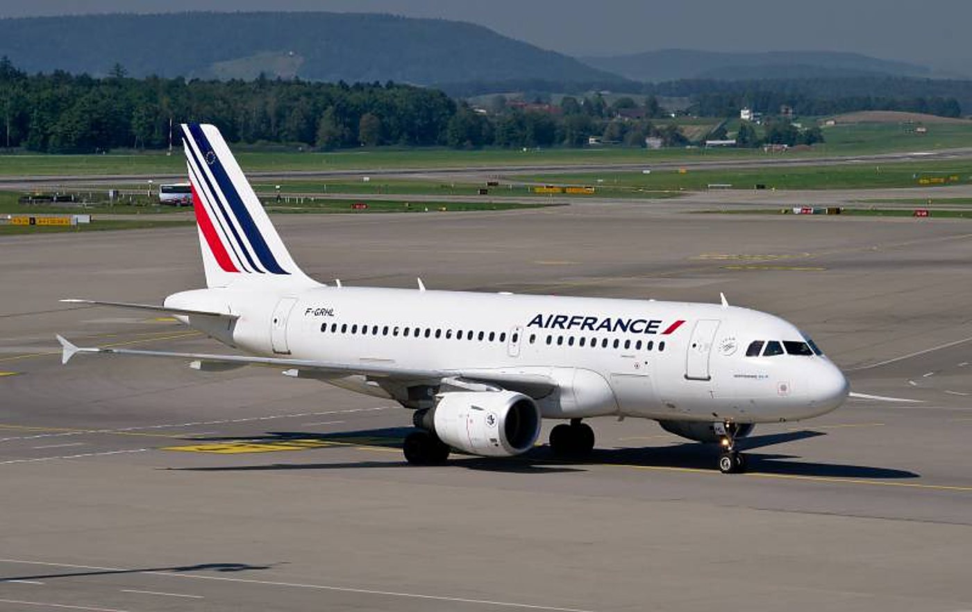 Un avion de ligne blanc aux couleurs d'Air France roule sur une piste d'aéroport, avec des collines boisées en arrière-plan.