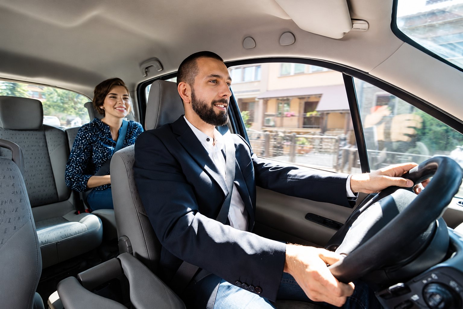 Un chauffeur barbu en costume sombre conduit une voiture tandis qu'une jeune passagère sourit sur la banquette arrière.