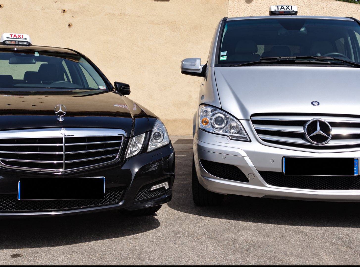 Une berline Mercedes noire et un monospace Mercedes gris, tous deux équipés d'un lumineux de toit taxi, garés côte à côte devant un mur beige.