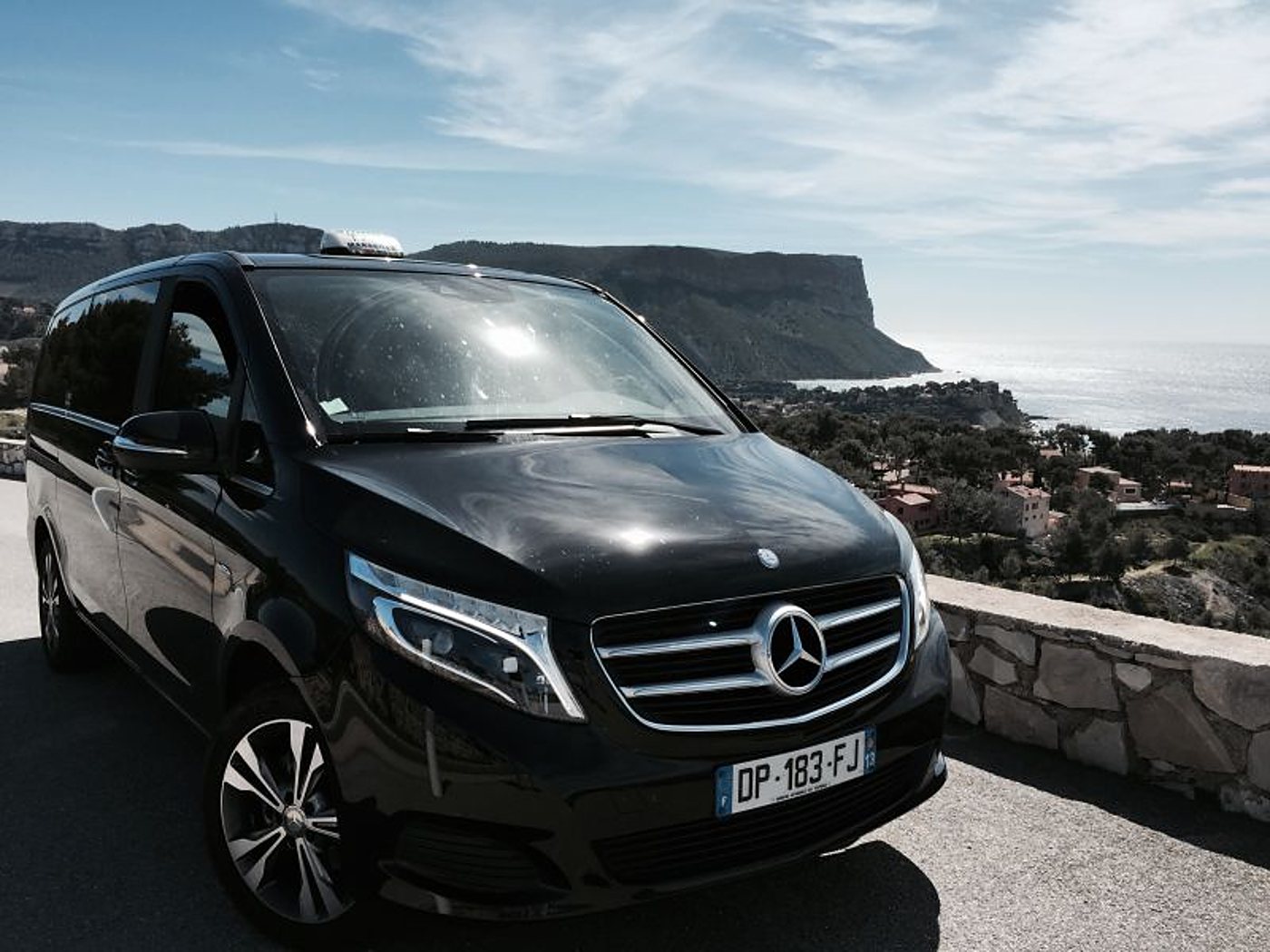 Un monospace Mercedes noir avec un lumineux de toit est garé sur une route en corniche, avec une falaise et la mer méditerranéenne en arrière-plan.