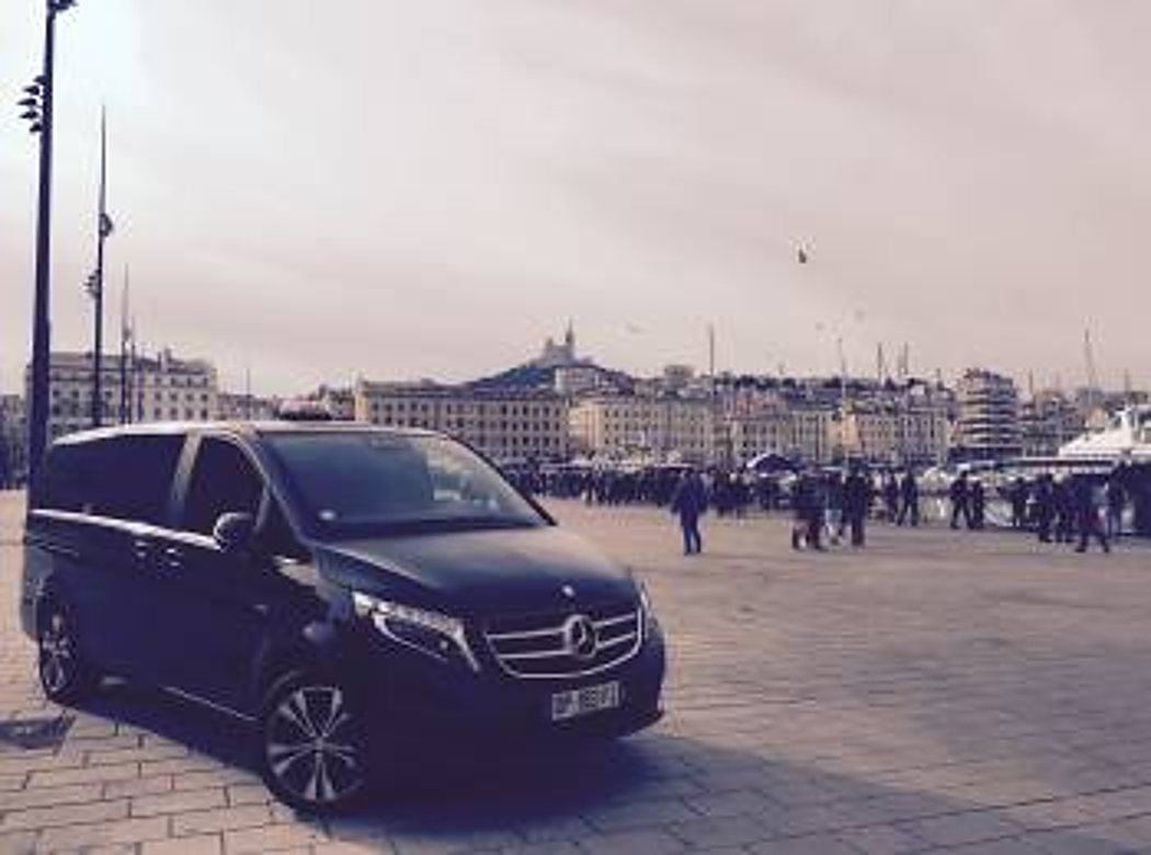 Un monospace Mercedes noir est garé sur un quai pavé face au port, avec des immeubles et des passants en arrière-plan sous un ciel nuageux.