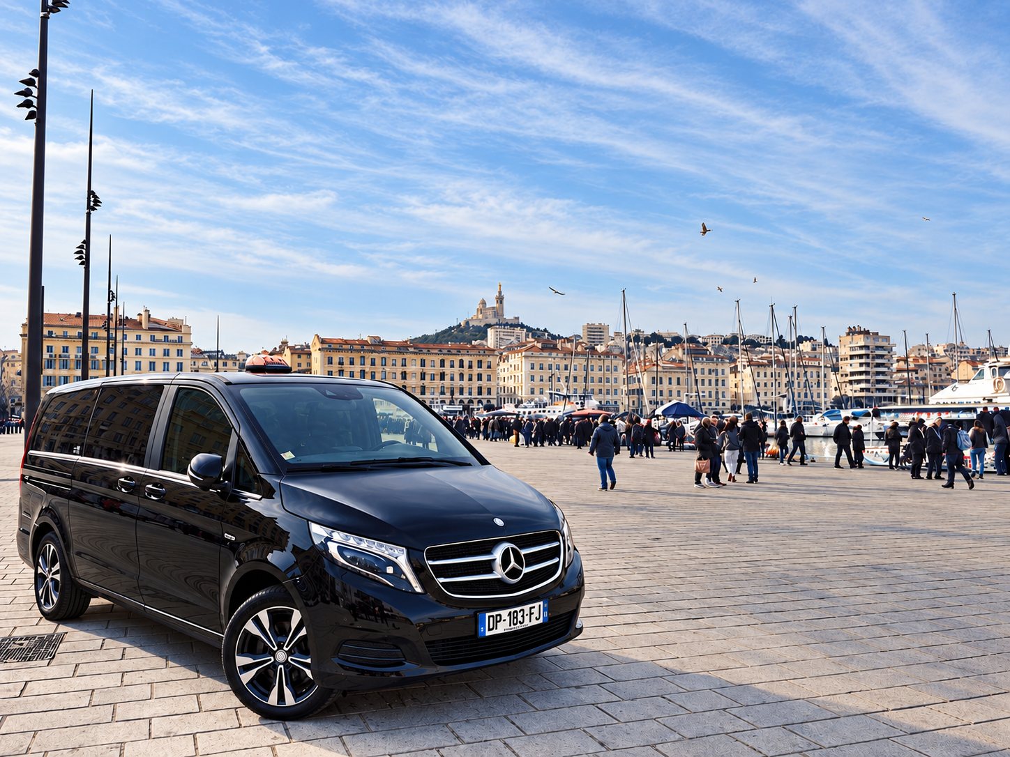 Minibus Mercedes noir garé sur les pavés du Vieux-Port de Marseille, la basilique Notre-Dame-de-la-Garde en arrière-plan.