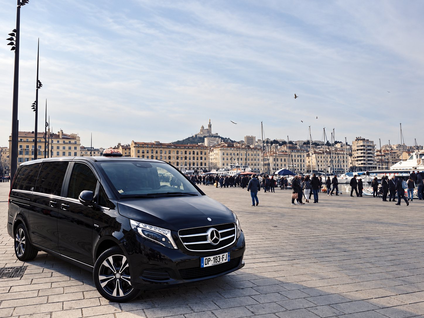 Un minibus Mercedes noir de type taxi est garé sur le quai du Vieux-Port, avec des passants, des bateaux et la basilique Notre-Dame-de-la-Garde en arrière-plan.
