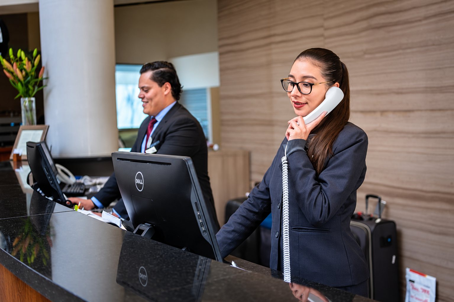 Une réceptionniste à lunettes téléphone au comptoir d'un hôtel pendant qu'un collègue en costume travaille derrière un écran.