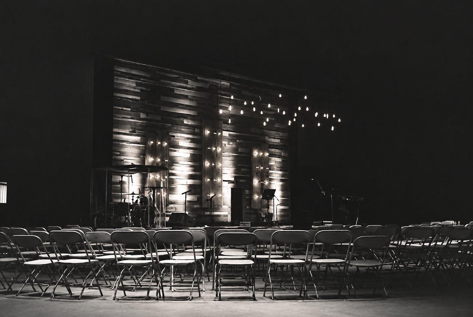 Photo en noir et blanc d'une salle vide où des rangées de chaises pliantes font face à une scène au fond de bois, éclairée par des guirlandes lumineuses et équipée d'une batterie et de micros.