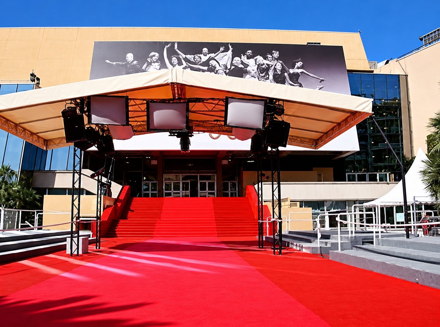 Un grand escalier recouvert de tapis rouge mène à l'entrée d'un palais des festivals surmonté d'une grande affiche en noir et blanc, sous un ciel bleu.