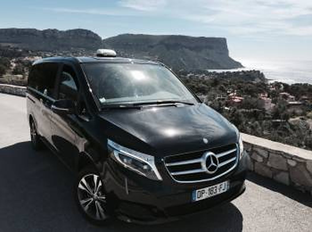 Un van Mercedes noir est garé sur une route en bord de mer, avec des falaises et la Méditerranée en arrière-plan.