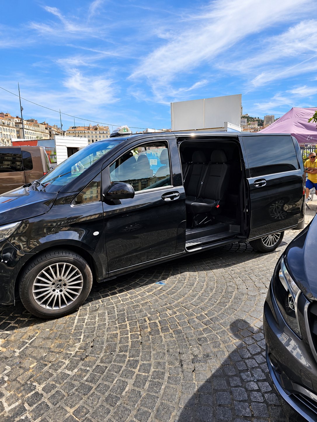 Un van noir avec une enseigne de taxi sur le toit est garé sur des pavés, sa porte coulissante ouverte laissant voir les sièges arrière.