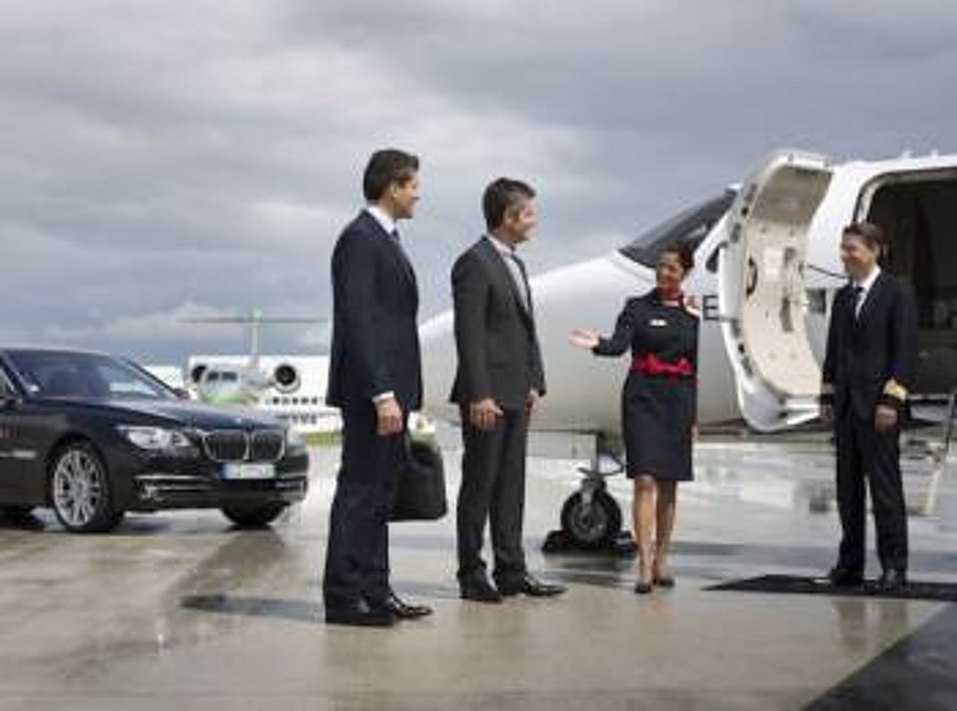 Deux hommes en costume sont accueillis par une hôtesse et un pilote devant un jet privé, à côté d'une berline noire sur le tarmac.